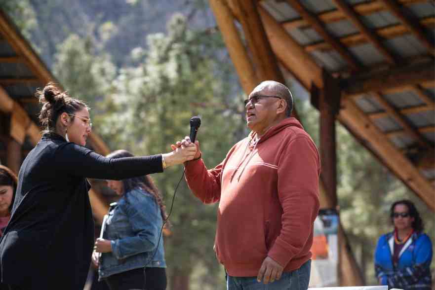 woman hands microphone to man in red shirt outside under wooden roof with trees in background