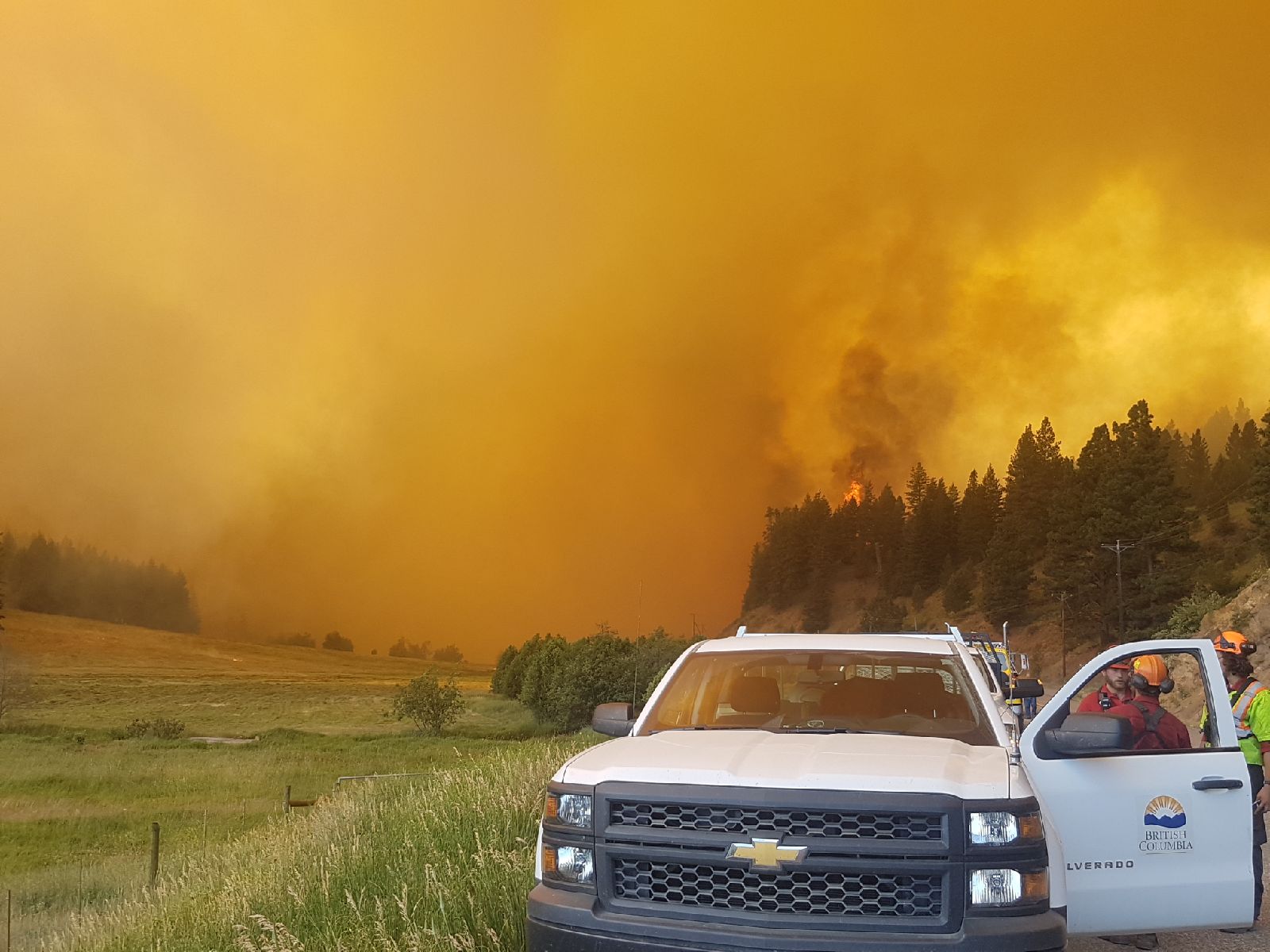 A BC government truck and wildfire personnel on a road with a wall of smoke and fire in the background