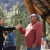 woman hands microphone to man in red shirt outside under wooden roof with trees in background